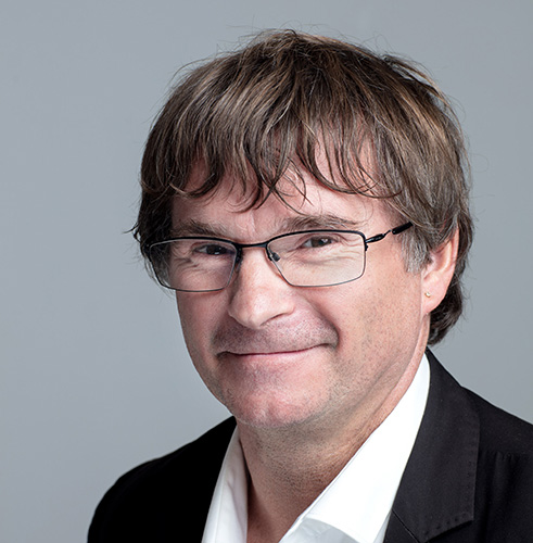 Portrait en studio d’un homme avec lunettes, cheveux bruns et veste de costume sombre sur chemise blanche, fond neutre