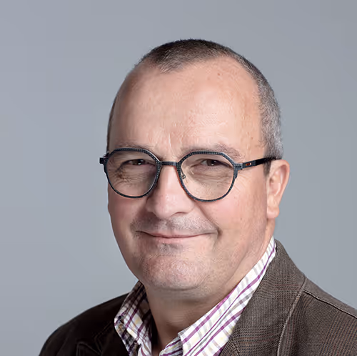 Portrait professionnel d’un homme souriant avec lunettes rondes, veste en velours et chemise à carreaux, sur fond uni gris