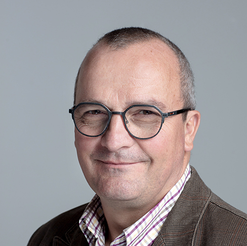 Portrait professionnel d’un homme souriant avec lunettes rondes, veste en velours et chemise à carreaux, sur fond uni gris