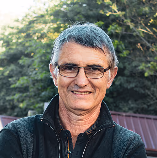 Portrait d’un homme en extérieur avec lunettes, cheveux gris et veste zippée, devant un fond de feuillage naturel