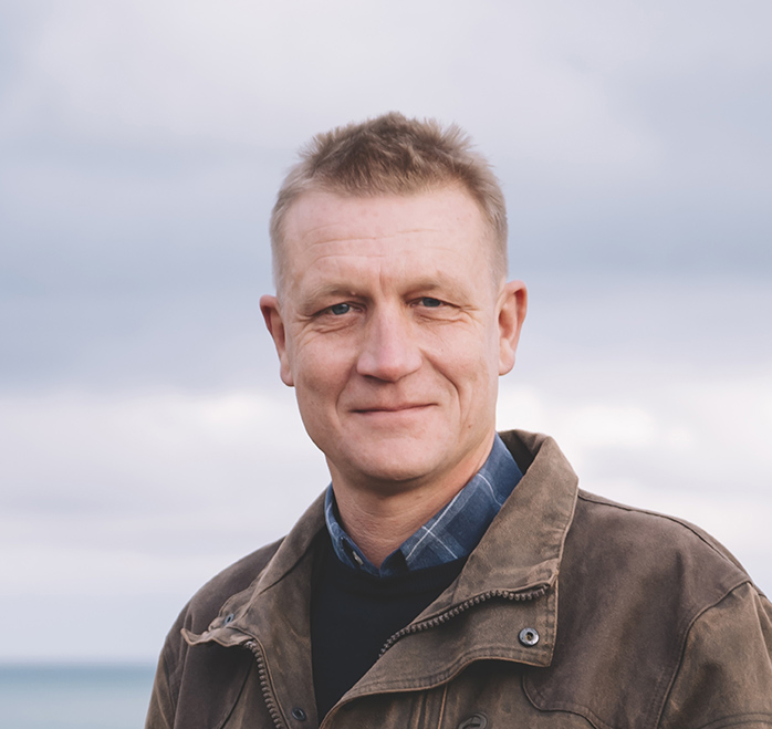 Portrait d'un homme en chemise bleue, en extérieur avec un paysage arboré en arrière-plan sous un ciel nuageux. Agriculteur debout devant un tas de betteraves en bordure d’un champ cultivé, sous un ciel nuageux