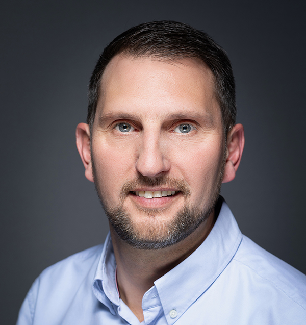 Portrait d’un homme souriant en chemise bleu clair, pris en studio sur fond sombre. Image adaptée à une présentation professionnelle ou institutionnelle.