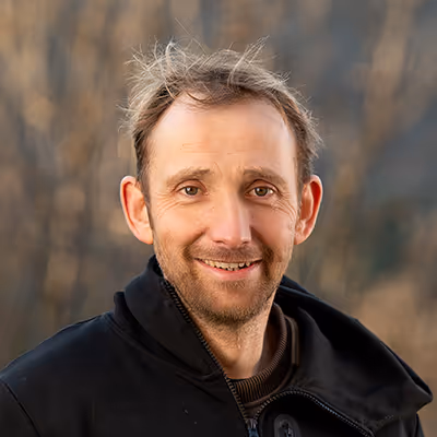 Portrait d’un homme souriant en extérieur, portant une veste noire, sur fond naturel légèrement flou.