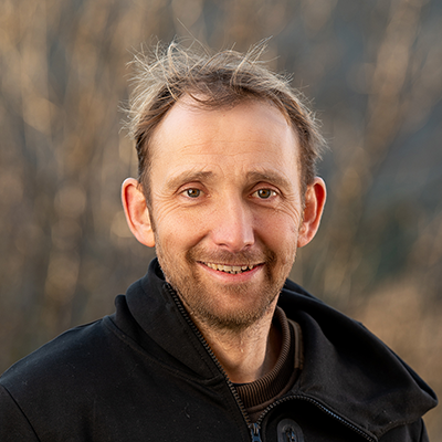 Portrait d’un homme souriant en extérieur, portant une veste noire, sur fond naturel légèrement flou.