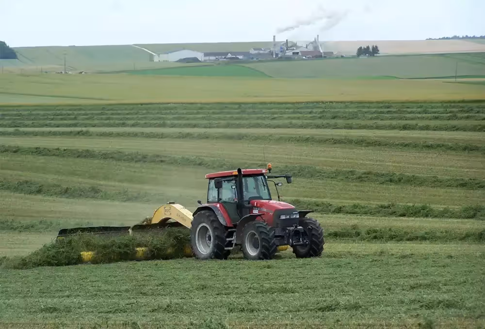 Tracteur rouge récoltant de l’herbe dans un champ verdoyant, avec des bâtiments industriels et de la fumée en arrière-plan.