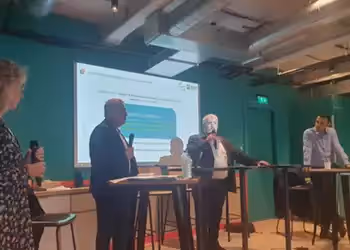 Présentation dans une salle avec des intervenants debout, échangeant avec le public devant un écran de projection.