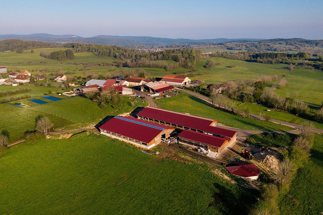 Vue aérienne d'une exploitation agricole avec plusieurs bâtiments aux toits rouges entourés de prairies verdoyantes et de collines boisées à l'horizon.