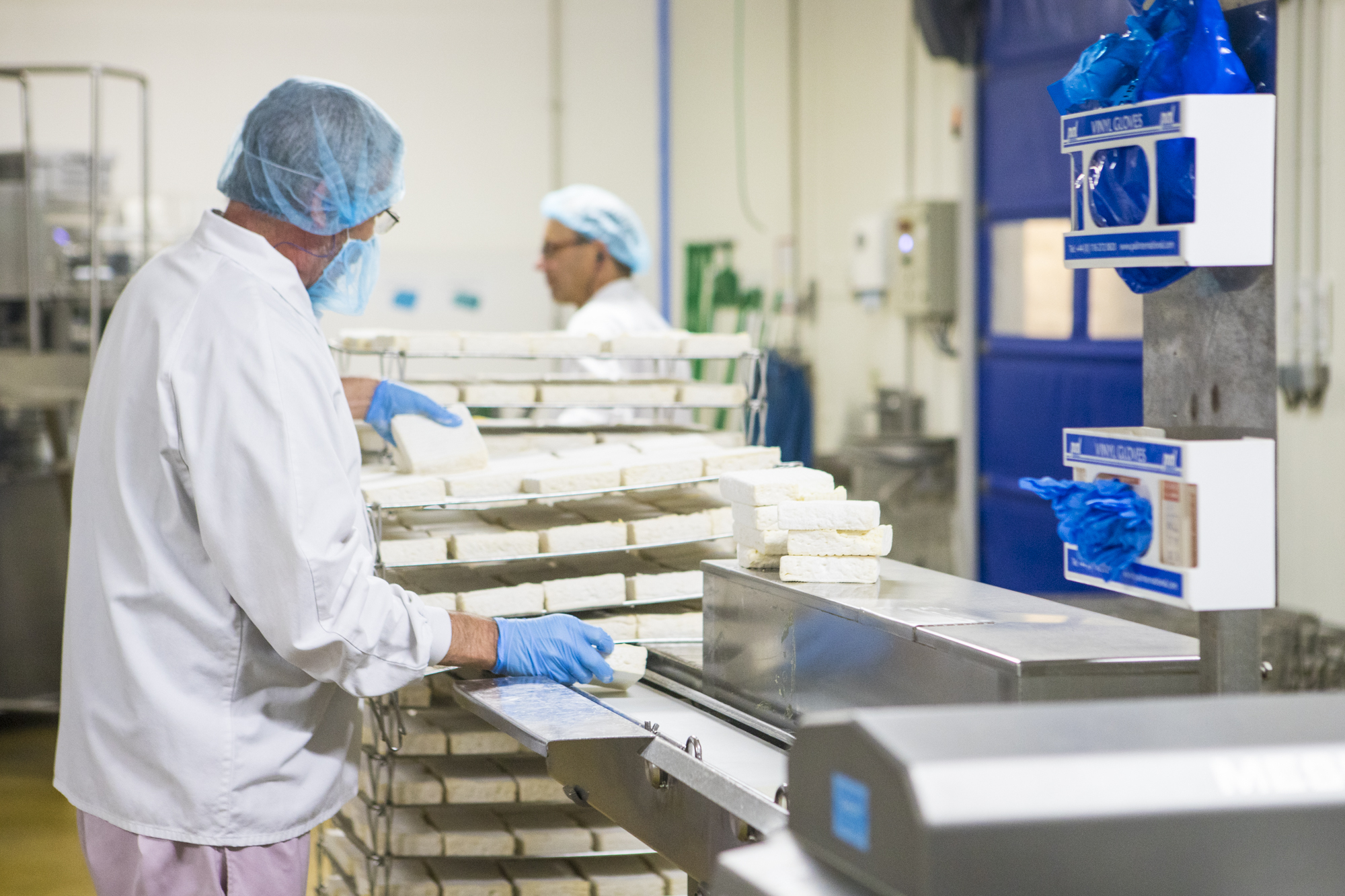 Travailleur en blouse blanche et charlotte manipulant des blocs de fromage sur une ligne de production dans une usine agroalimentaire, illustrant le processus de transformation fromagère en environnement contrôlé.