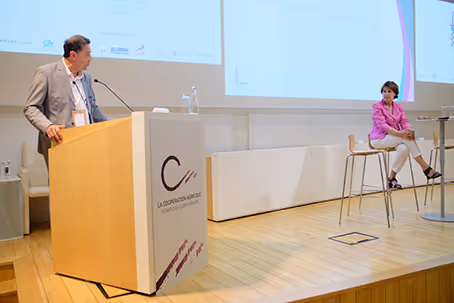 Conférence sur la coopération agricole avec un intervenant à la tribune et une intervenante assise sur scène.