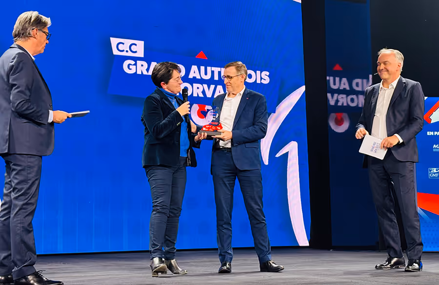 Remise de prix sur scène lors du Grand Prix des Maires 2025, avec quatre personnes en costume, dont une femme remettant un trophée à un lauréat, sur fond d’écran bleu affichant le nom de la communauté de communes « Grand Autunois Morvan ».