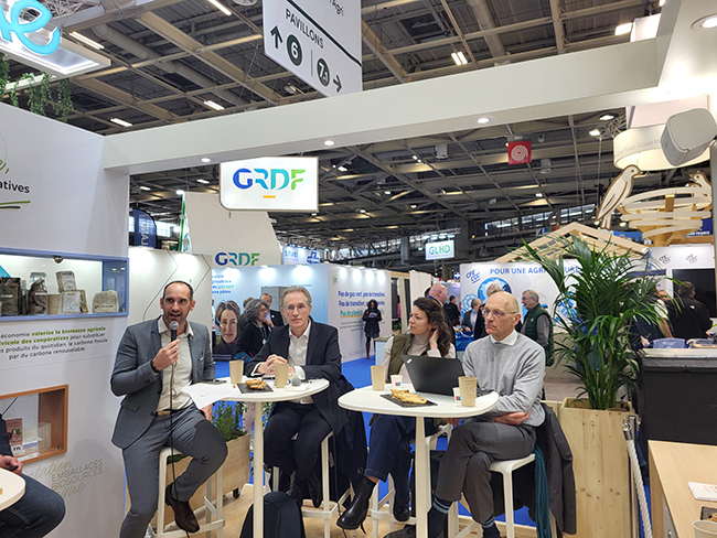 Table ronde organisée sur un salon professionnel, avec plusieurs intervenants assis autour de tables hautes, échangeant sur la bioéconomie devant le stand de GRDF.