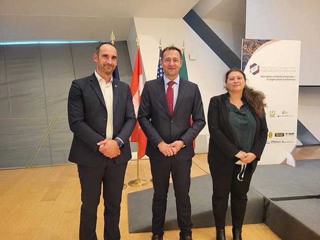 Photo officielle lors d’une conférence agricole internationale, avec trois intervenants posant devant les drapeaux de plusieurs pays et un kakémono de l'événement.