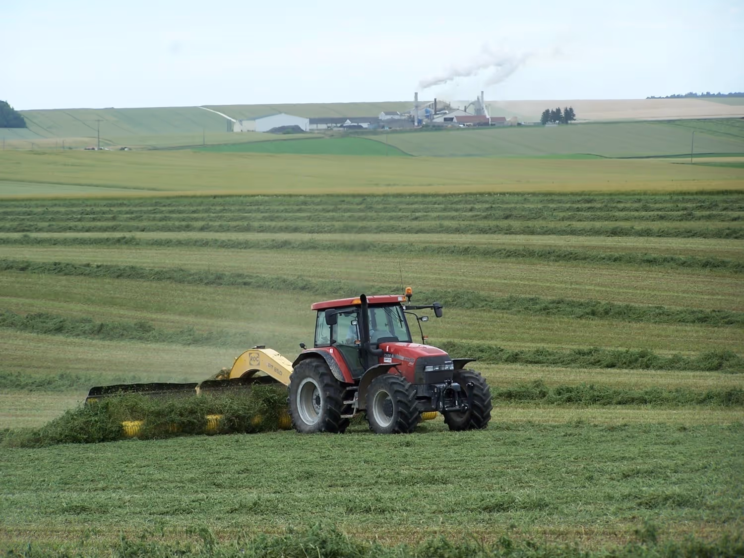 Tracteur rouge récoltant de l’herbe dans un champ verdoyant, avec des bâtiments industriels et de la fumée en arrière-plan.