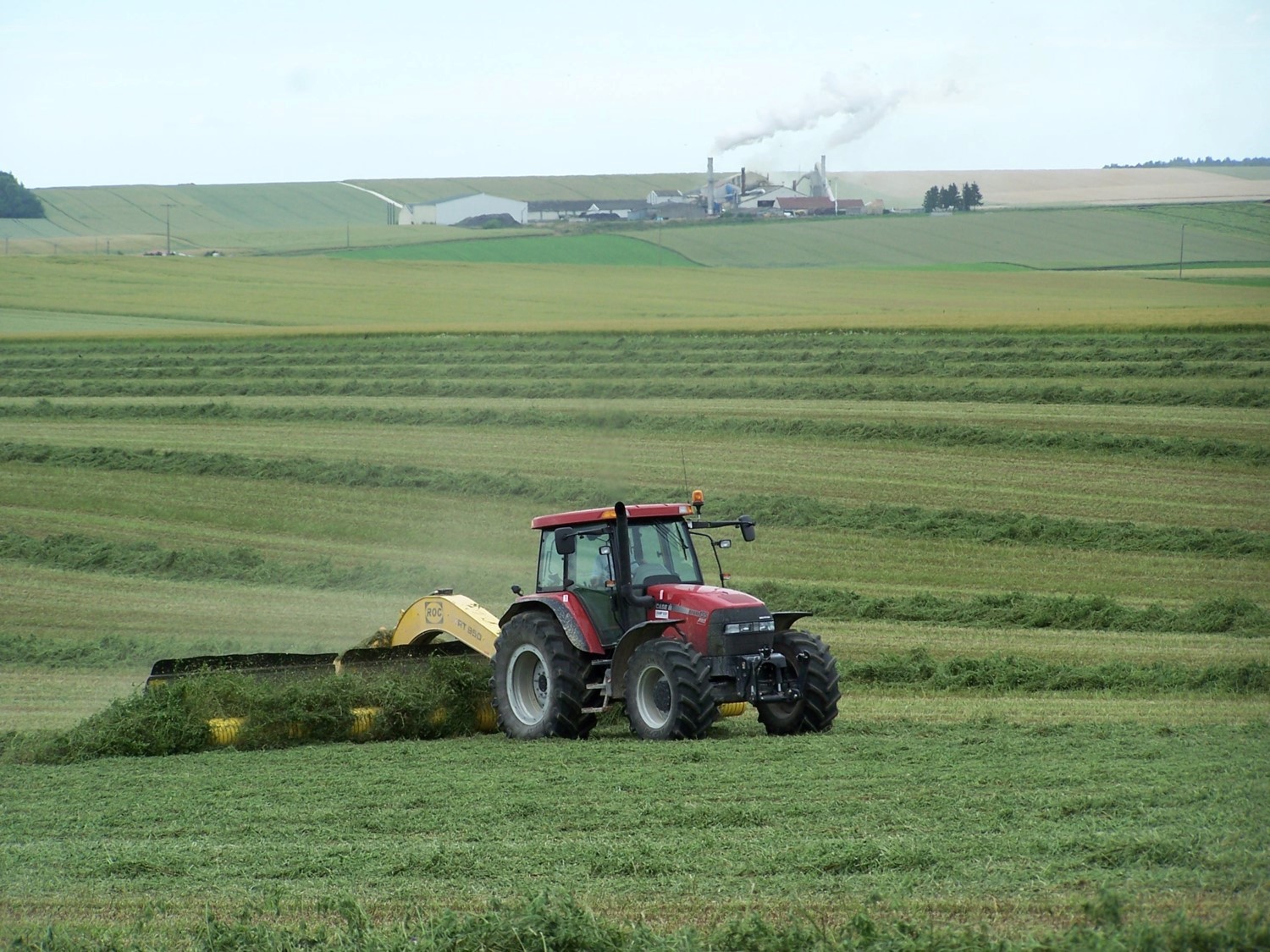 Tracteur rouge récoltant de l’herbe dans un champ verdoyant, avec des bâtiments industriels et de la fumée en arrière-plan.