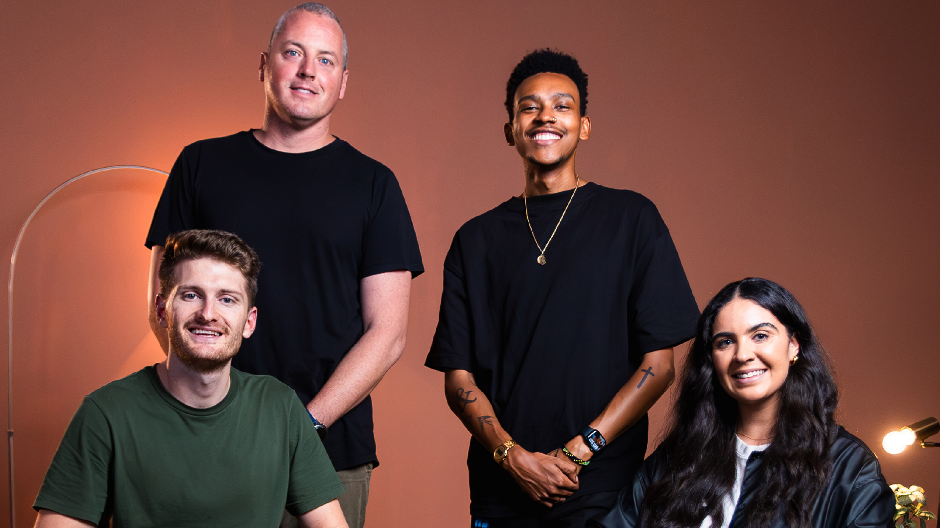 Group of four diverse young adults smiling and posing against a plain brown background with soft lighting.