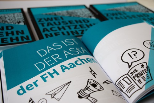 Aufgeschlagener Kalender mit Text und Illustrationen, davor liegen weitere gefaltete Exemplare mit blauer Titelseite.