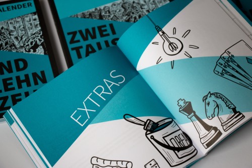 Aufgeklappter Kalender mit der Überschrift ‚EXTRAS‘ und Illustrationen wie Glühbirne, Farbeimer, Schachfiguren und Fächer.
