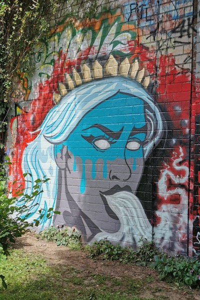 Wandgemälde einer blauen Frau mit goldener Krone, die aus dem Mund Rauch speit, auf einer Backsteinwand mit rotem und buntem Graffiti.