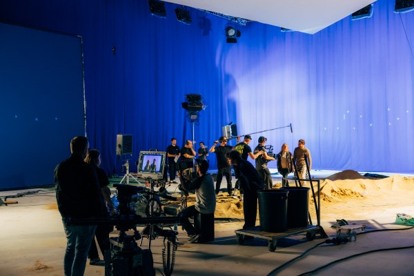 Filmcrew bei der Aufnahme in einem Studio mit blauem Hintergrund und künstlichem Sand auf dem Boden.