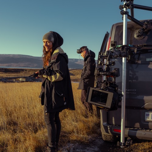 Nikita steht lächelnd in einem Feld, während ein Mann mit VR-Brille neben einem Fahrzeug mit montierter Kameratechnik steht.