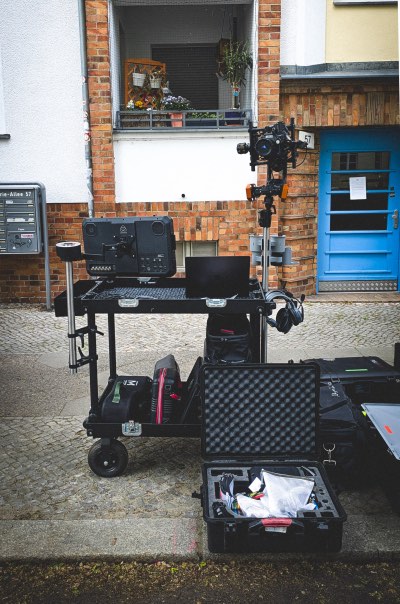 Kameraausrüstung mit Stativ, Laptop und geöffnetem Koffer auf einem Rollwagen vor einem Backsteinhaus mit blauem Tür.