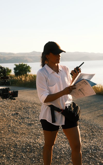 Nikita im Freien bei Sonnenuntergang trägt weiße Bluse, schwarze Shorts und Kappe, hält Walkie-Talkie und Papiere in der Hand.