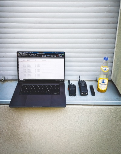 Offener Laptop mit einem Dokument auf dem Bildschirm, daneben zwei Funkgeräte, ein USB-Stick und eine Flasche Club-Mate auf einem Fensterbrett.