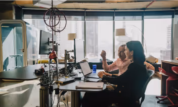 Two people working on laptops at a desk in a modern office with large windows and city views.