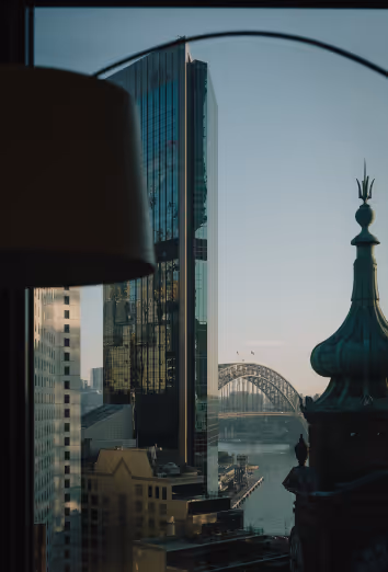 View of a city skyline with a reflective glass skyscraper, a lit building with a green rooftop spire, and a distant arched bridge over water at sunset.