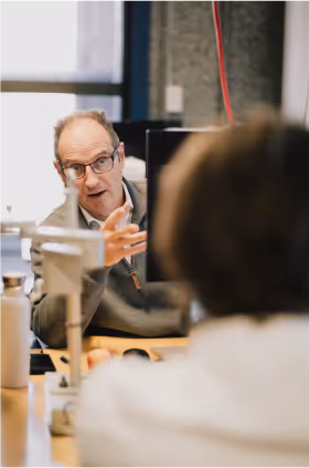 Man with glasses gesturing while talking to another person in an office setting.