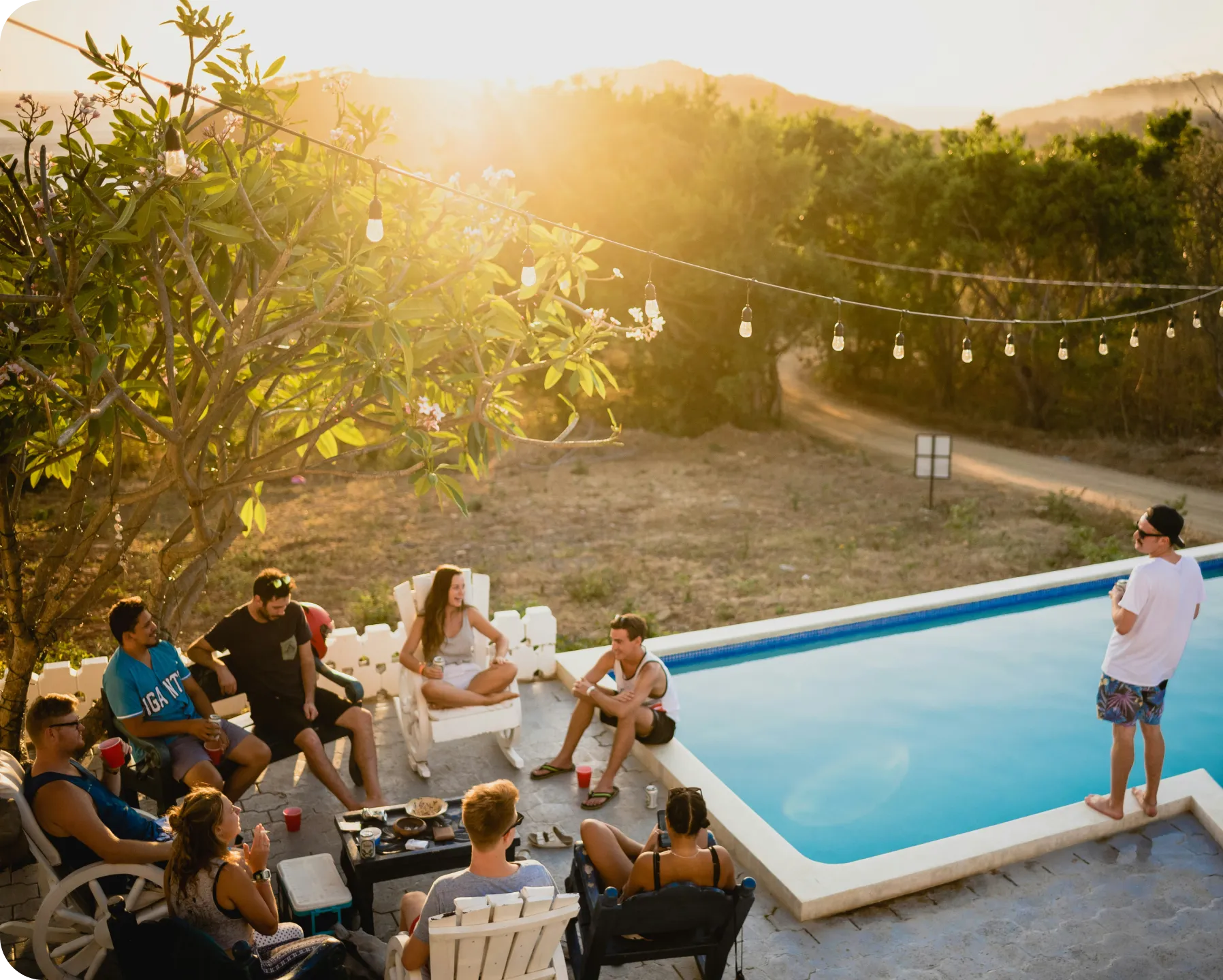 Group of friends socializing and relaxing by a pool at sunset with string lights overhead.