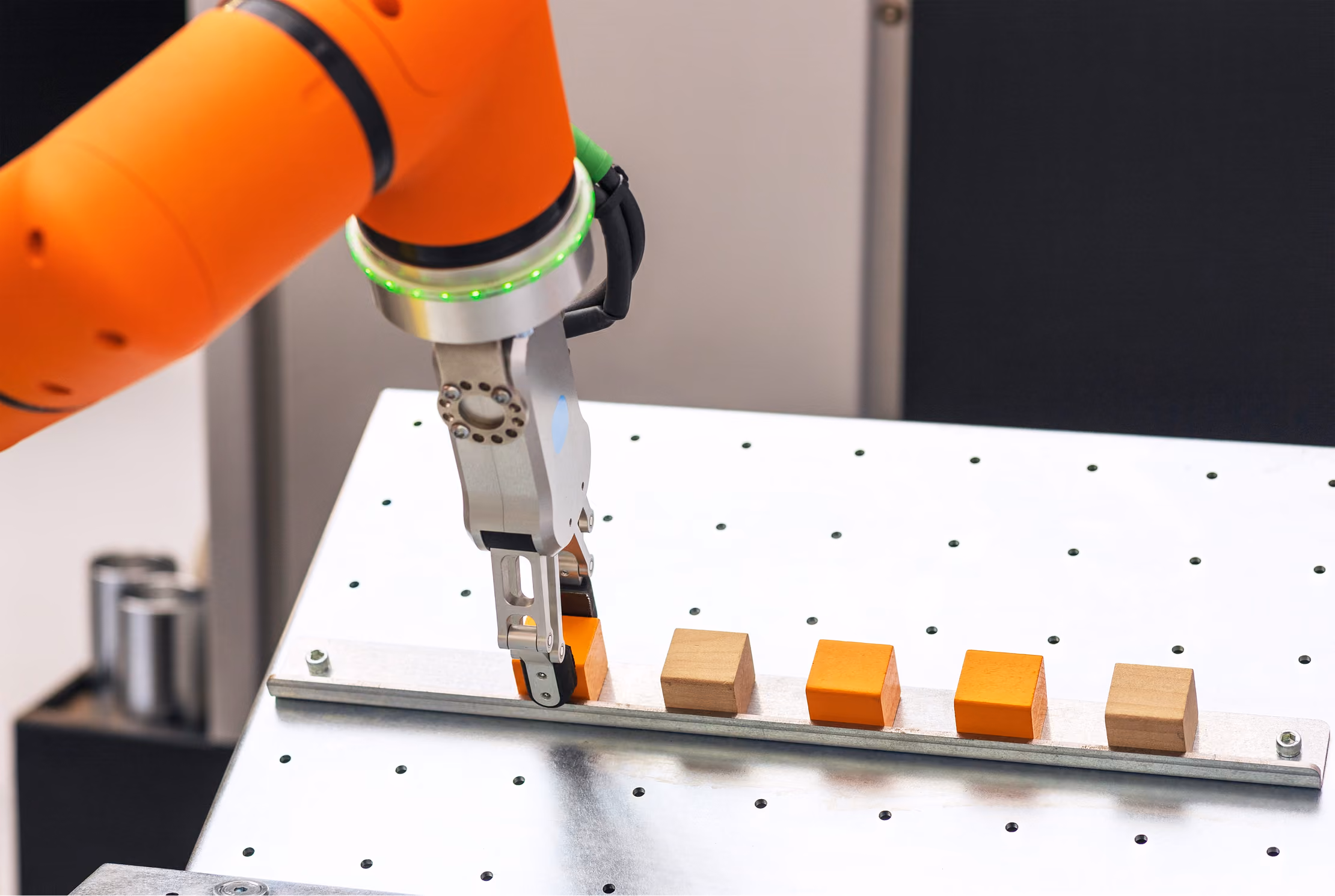 Bras robotisé orange saisissant un cube orange sur une plaque métallique percée avec plusieurs cubes en bois et orange alignés.