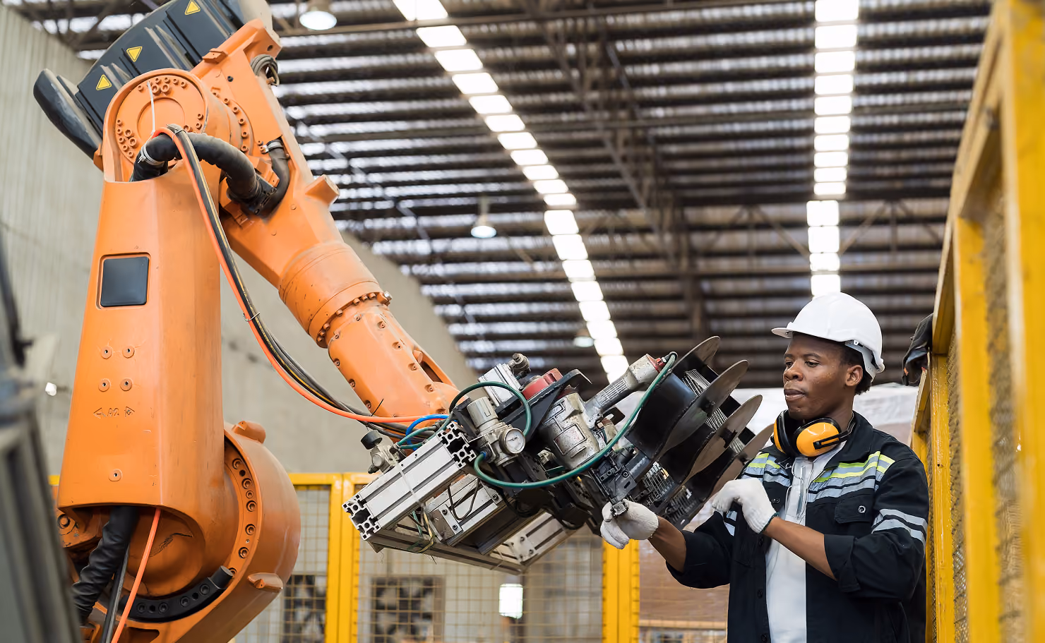 Un ouvrier portant un casque blanc, des protège-oreilles et des gants ajuste un bras robotique orange dans un grand atelier industriel.