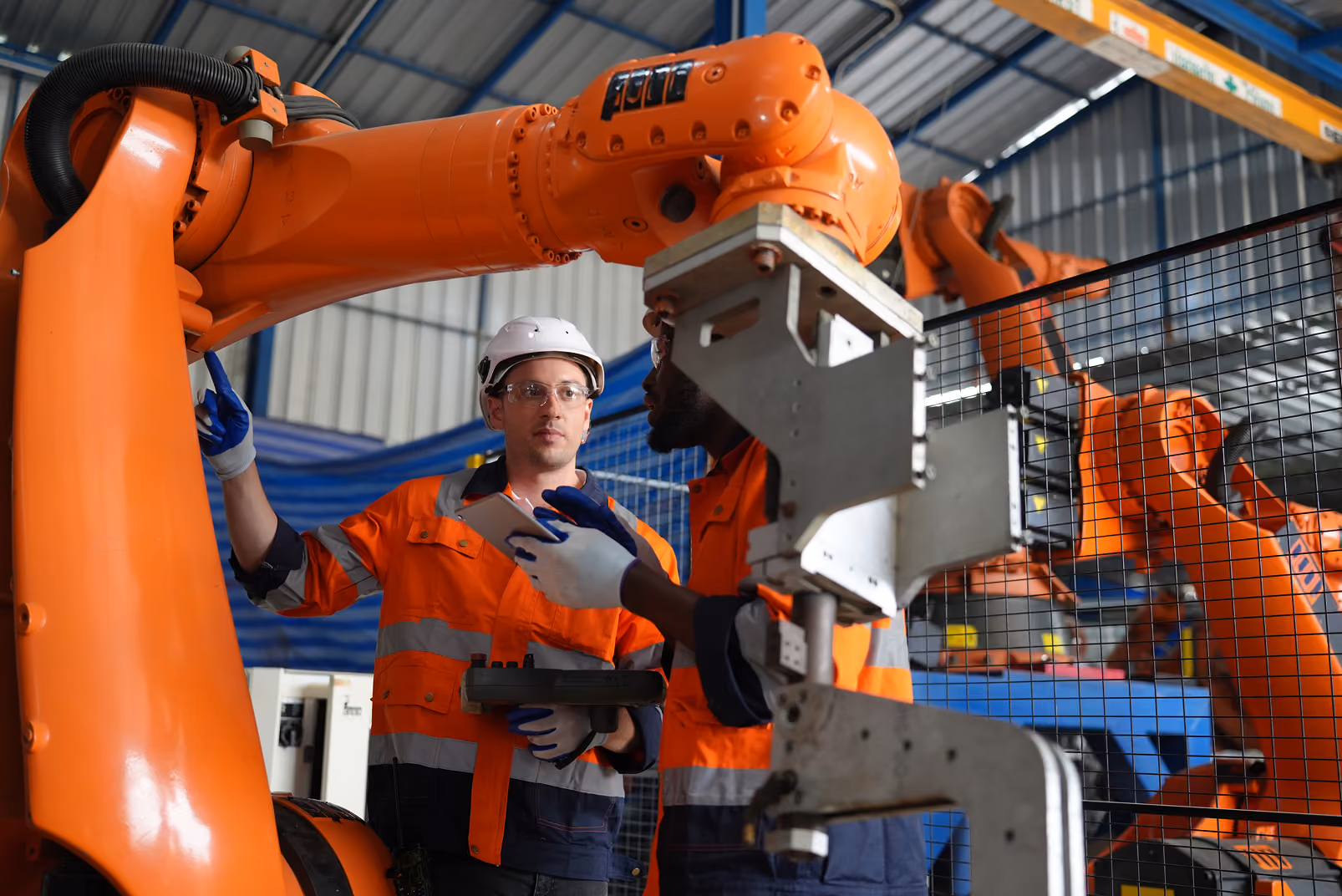 Deux ouvriers en tenue de sécurité orange avec casques et lunettes discutent devant des bras robotisés orange dans une usine.