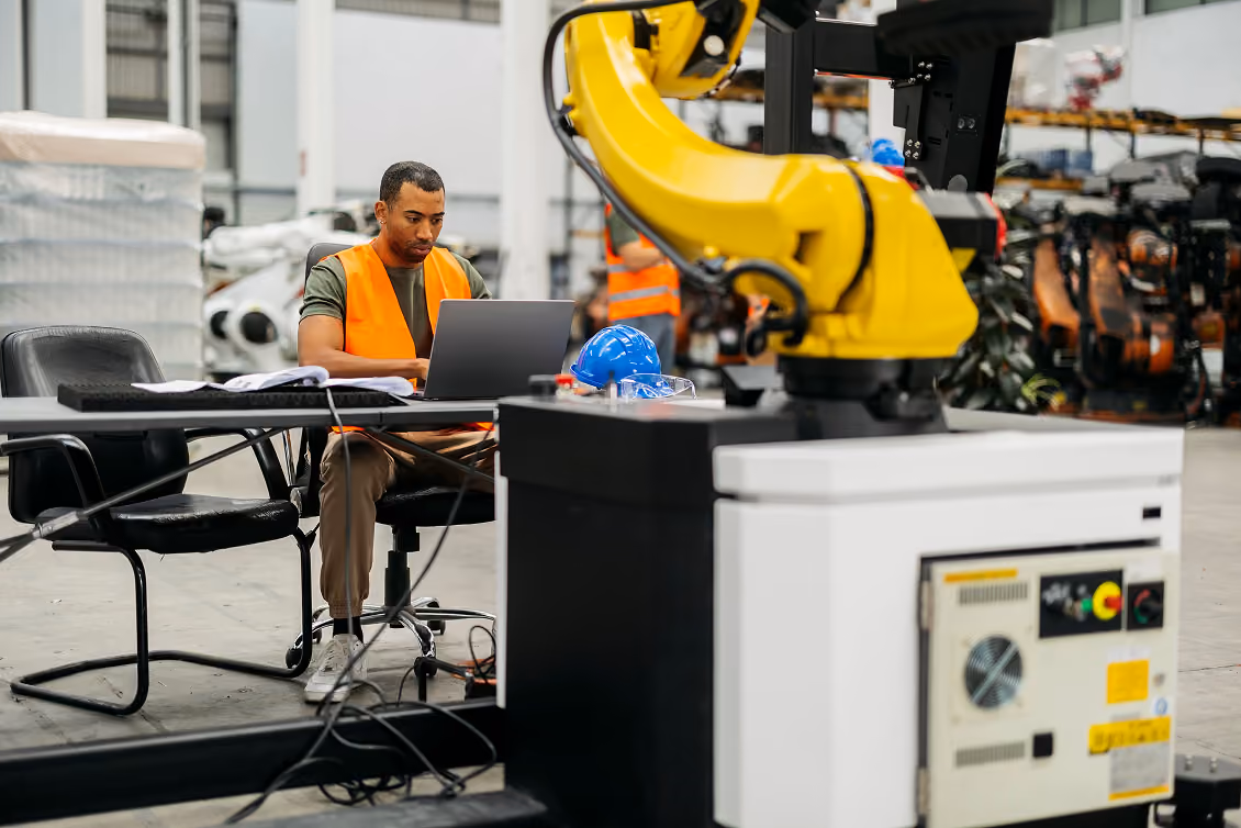 Un homme portant un gilet de sécurité orange utilise un ordinateur portable assis à une table dans un atelier industriel avec un robot jaune en premier plan.