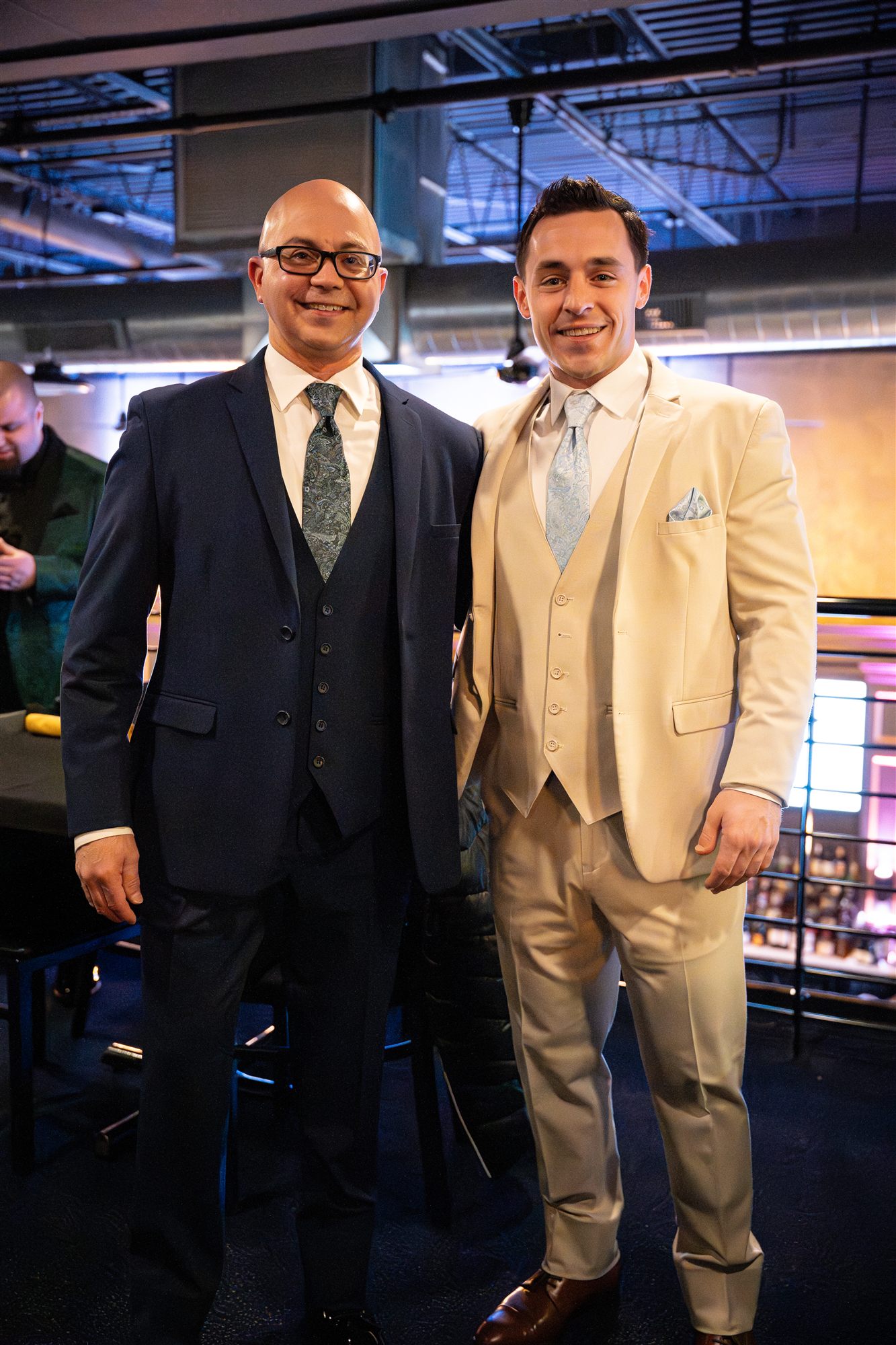 Two men in sharp suits posing arm-to-arm on the mezzanine in warm stage light