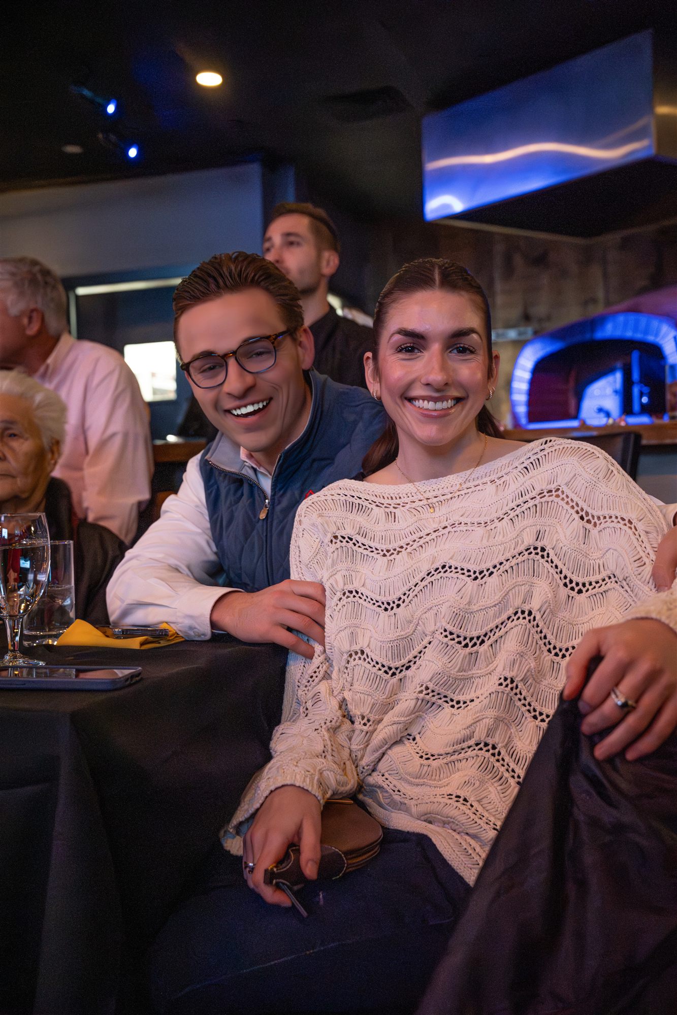 Smiling couple seated close at a black-linen table by the wood-fired oven