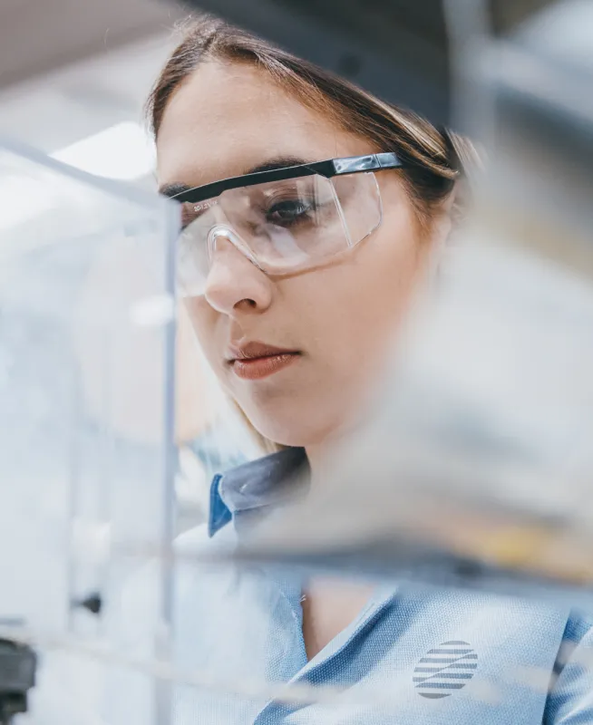 Woman wearing safety glasses focused on a clear plastic surface in a laboratory or work setting.