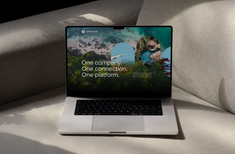 Open laptop on a beige fabric sofa displaying a travel-themed screen with coastal and tropical images and text reading 'One company. One connection. One platform.'