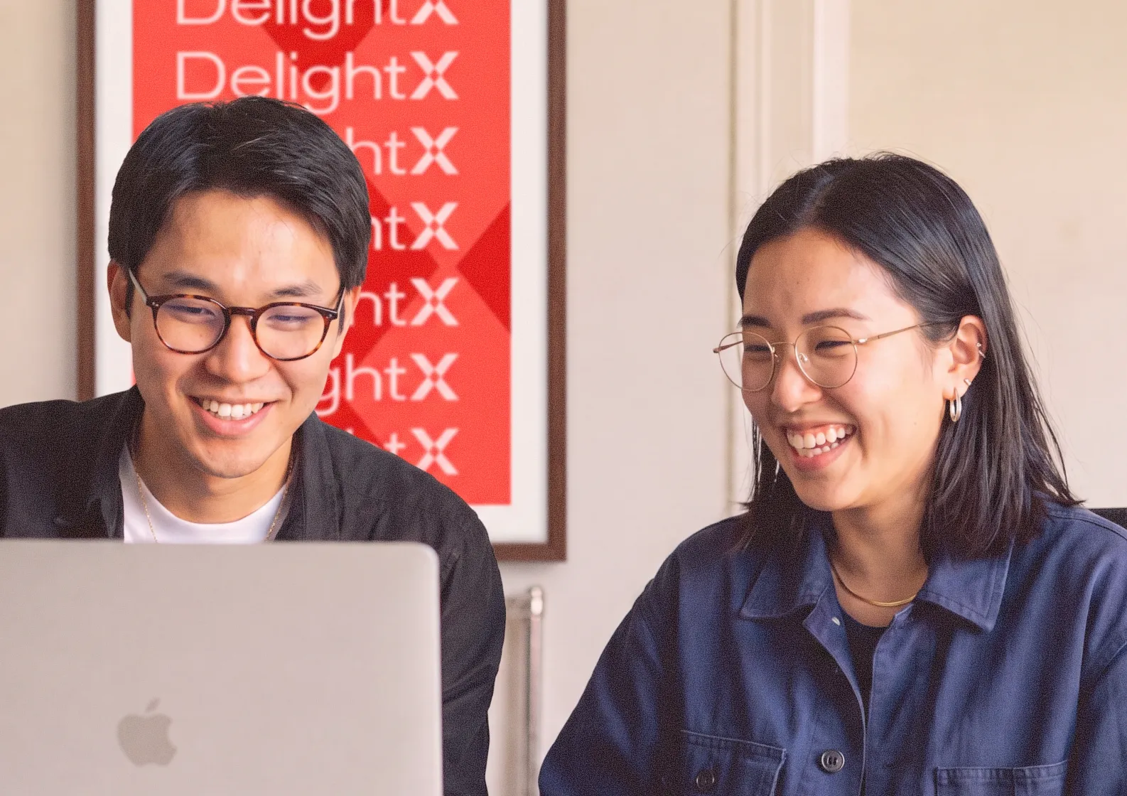 Two smiling people with glasses looking at a laptop in front of a red DelightX sign.