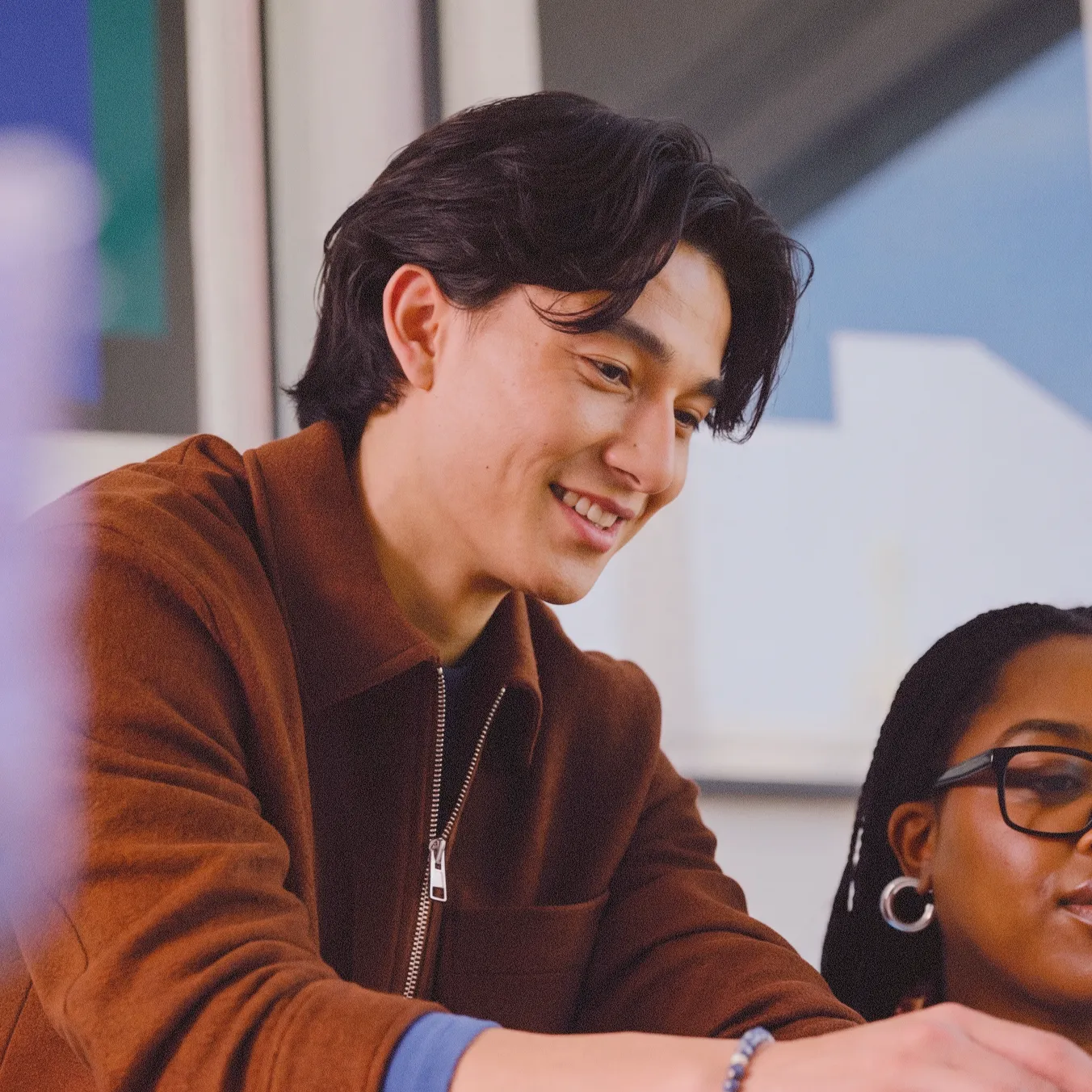 Smiling young man in brown jacket looking down, next to a woman wearing glasses and hoop earrings.