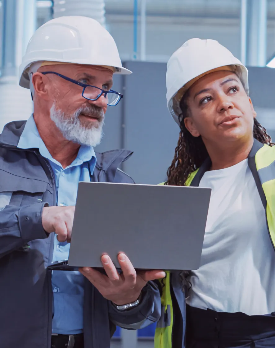 Two engineers wearing hard hats checking a laptop in an industrial setting.