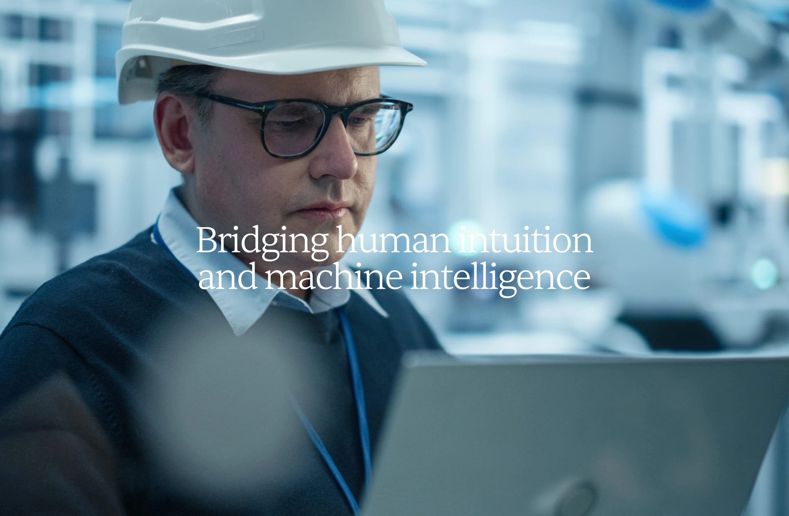 Man wearing a hard hat and glasses working on a laptop in an industrial setting.
