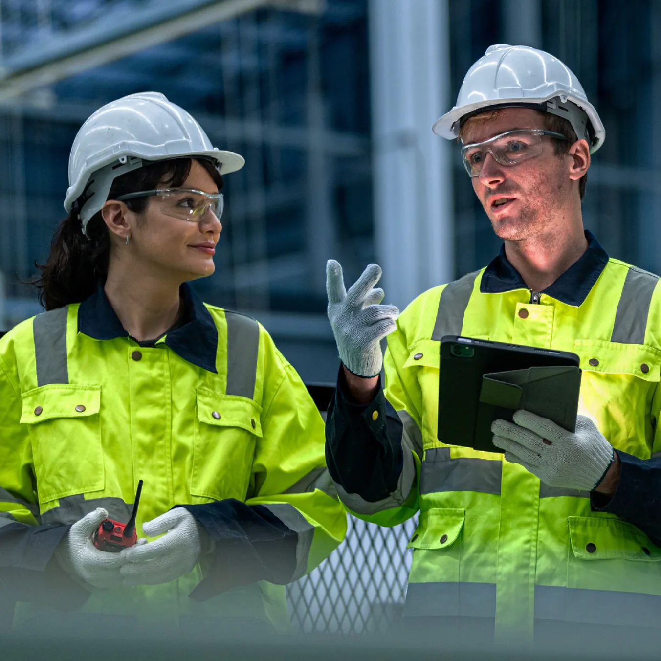 Two construction workers in safety gear discussing while holding a walkie-talkie and tablet indoors.