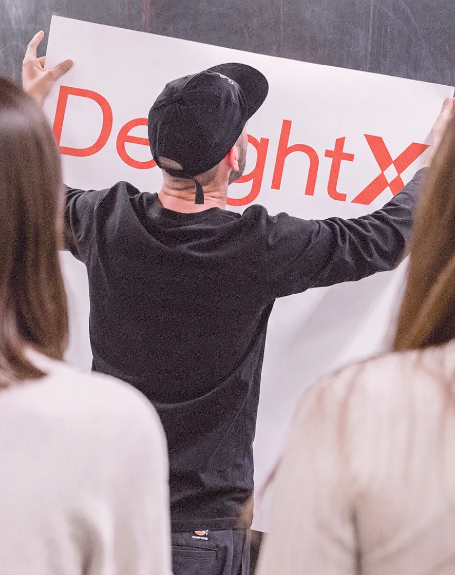 Man in black cap placing a poster with the word 'DeightX' in front of a chalkboard while two women watch.