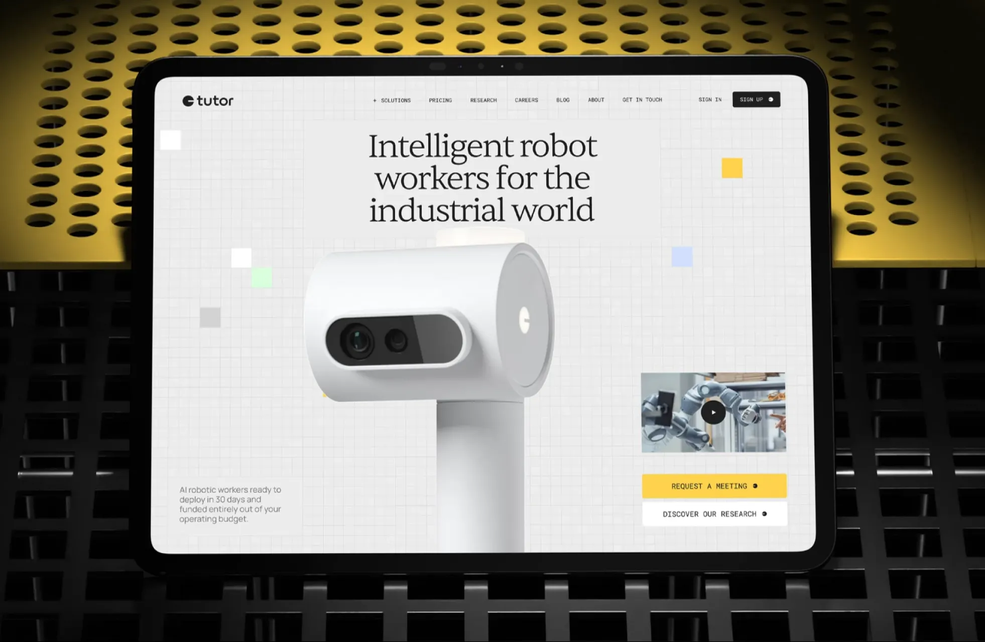 Tablet screen displaying a webpage for Tutor Intelligence showing a white robotic camera with two lenses, text stating 'Intelligent robot workers for the industrial world,' a video thumbnail of robotic arms, and buttons labeled 'Request a Meeting' and 'Discover Our Research.'