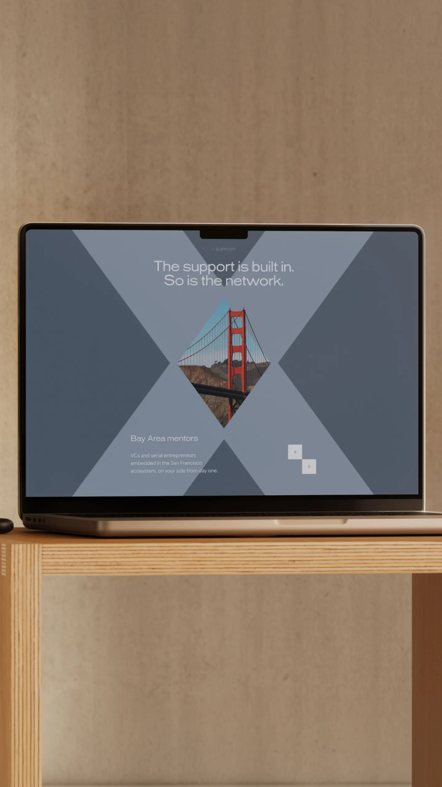 Laptop on a wooden table displaying a webpage with a large X design framing a diamond-shaped photo of the Golden Gate Bridge and text saying 'The support is built in. So is the network.' and 'Bay Area mentors' with a description about VCs and serial entrepreneurs in the San Francisco ecosystem.