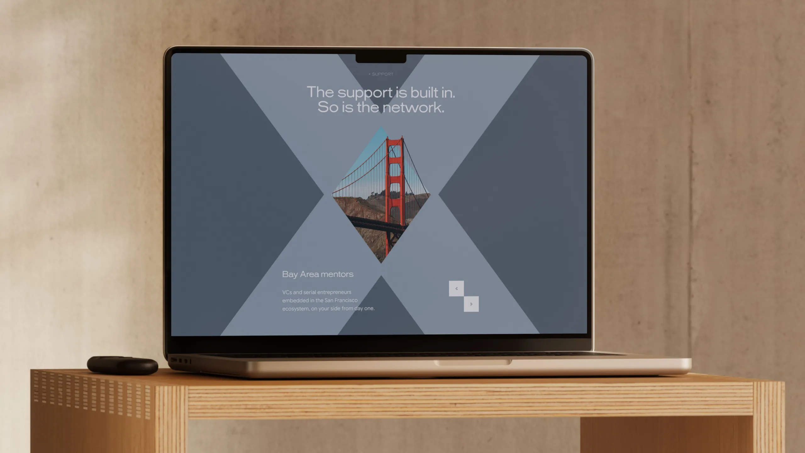 Laptop on a wooden table displaying a digital design with a diamond-shaped image of the Golden Gate Bridge and text reading 'The support is built in. So is the network.' with a description about Bay Area mentors and San Francisco ecosystem.