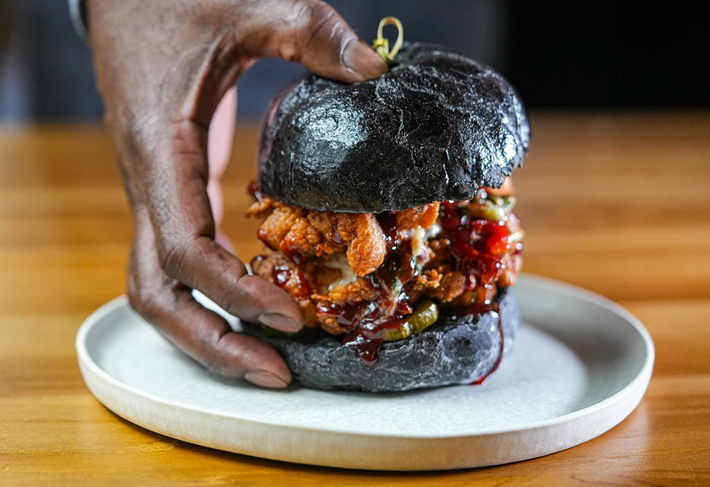 Hand holding a fried chicken sandwich with black bun and barbecue sauce on a white plate.
