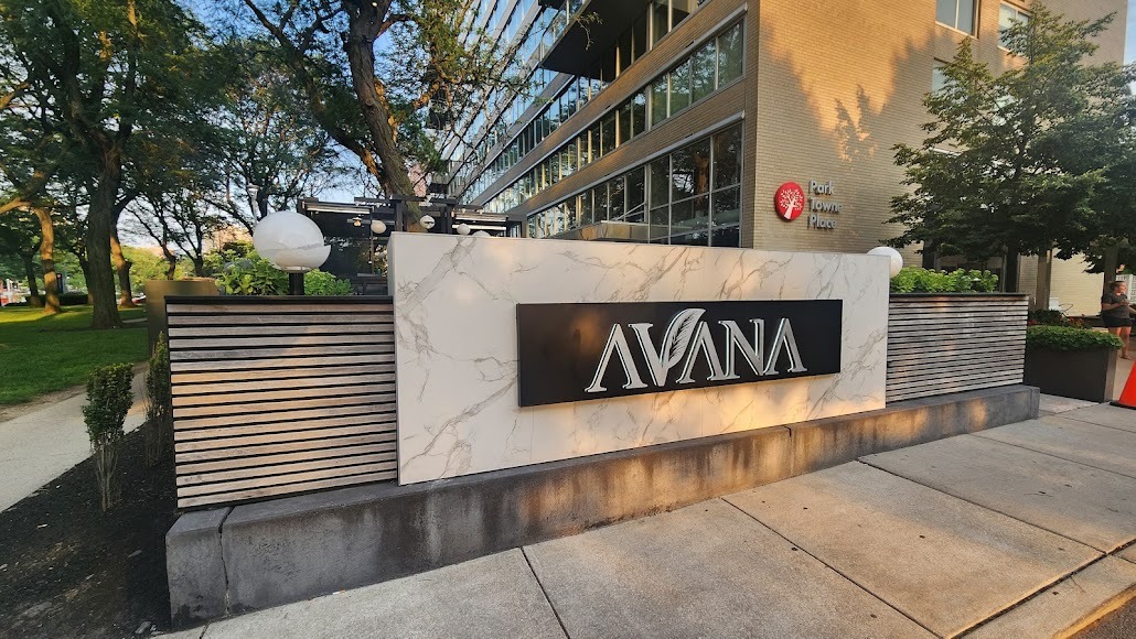 Outdoor sign of the restaurant 'AVANA' mounted on a marble-patterned panel in front of a modern building with trees around.